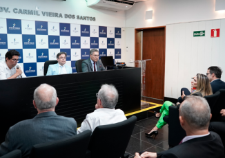 Há três homens sentados. Eles estão de frente a uma plateia que asssite um dos homens falando.