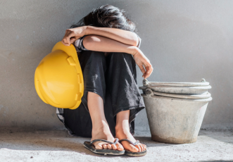 Criança sentada no chão com as costas apoiadas em uma parede. Ela usa calça preta e está com as duas pernas dobradas e os dois braços por cima dos joelhos cobrindo o rosto e segurando um capacete amarelo com a mão esquerda