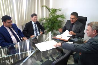 A imagem mostra quatro homens sentados ao redor de uma mesa de vidro em uma reunião. Eles estão vestidos formalmente, usando ternos e gravatas. O homem ao centro, que parece estar conduzindo a reunião, veste um terno escuro e óculos, segurando um papel em suas mãos. À sua direita, um homem de terno azul e gravata listrada tem uma expressão séria, enquanto outro homem ao lado dele, de terno preto e gravata listrada, sorri levemente. 