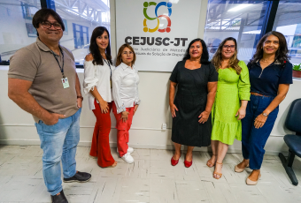 Cinco mulheres e um homem em pé na frente de uma placa com a palavra Cejusc-JT. 