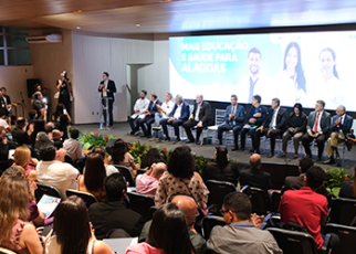 A imagem mostra um evento em um auditório, com um grande público acompanhando uma solenidade. No palco, diversas autoridades estão sentadas em uma mesa de honra, enquanto uma pessoa discursa em pé ao lado. Ao fundo, um telão exibe a mensagem "Mais Educação e Saúde para Alagoas", indicando o tema da cerimônia. O ambiente está decorado com arranjos florais na frente do palco, e a plateia acompanha atentamente.