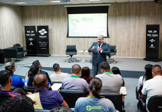 Há um homem de terno cinza falando de pé para uma plateia senda em um auditório