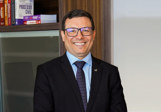 Homem de pele clara, cabelos curtos e escuros, usando óculos e sorrindo. Ele veste terno preto, camisa azul clara e gravata azul escura com pequenos pontos. Ao fundo, há uma estante com livros jurídicos.