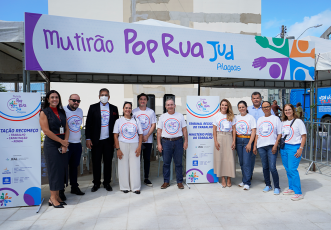 Há 11 pessoas em pé, sorrindo, em frente a uma tenda com uma grande faixa escrita "Mutirão Pop Rua Jud Alagoas". A maioria das pessoas veste camisetas brancas do evento, com o logotipo colorido do “Pop Rua Jud” no centro. Duas mulheres estão com roupas diferentes.