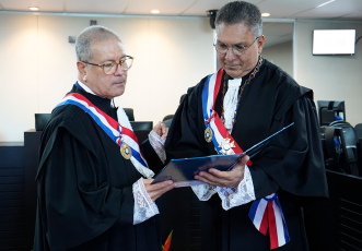 Dois magistrados estão em destaque, trajando togas pretas com detalhes em renda branca nos punhos e no colarinho, além de faixas nas cores azul, branca e vermelha, atravessadas no peito, e medalhas penduradas. Eles estão em pé dentro de uma sala de sessões, em um ambiente institucional, olhando atentamente para um documento azul que seguram juntos