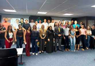 Fotografia mostra um grupo de pessoas reunidas em um auditório para uma foto coletiva. Elas estão dispostas em duas fileiras: a primeira em pé no chão do auditório e a segunda em pé sobre um pequeno palco. Ao fundo, há dois telões de projeção exibindo slides com rostos de outras pessoas, sugerindo uma apresentação.