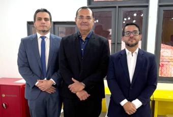 Três magistrados posam juntos para foto em ambiente interno, vestindo ternos formais. Ao fundo, janelas com cortinas translúcidas e mobiliário escolar.