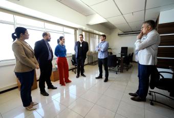 Há seis pessoas em pé em uma sala ampla e bem iluminada, conversando em roda. A cena ocorre em um ambiente corporativo, com mesas, cadeiras e janelas grandes ao fundo.