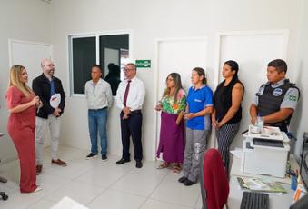 Grupo de oito pessoas está reunido em pé dentro de uma sala clara. Elas estão lado a lado, algumas em trajes formais e outras com roupas casuais. À direita da imagem, há um agente de segurnaça patrimonial fardado ao lado de uma mesa com computador e documentos.