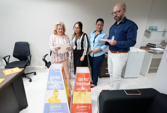 A foto mostra a desembargadora Vanda Lustosa e três integrantes do Subcomitê de Prevenção e Enfrentamento ao Assédio do TRT da 19ª Região durante a abertura das “Urnas do Desabafo”. Eles estão reunidos em uma sala, lendo os papéis recolhidos das urnas identificadas com as frases “Quebre o silêncio” e “Sua voz tem poder”, que incentivam o enfrentamento ao assédio no ambiente de trabalho.