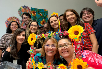 Foto de onze pessoas, sendo mulheres e quatro homens, sendo as mulheres vestidas de roupas juninas e com ornamentos nas mãos.