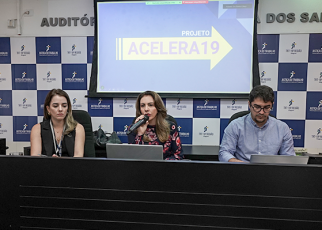 Foto de três pessoas sentadas à mesa, sendo duas mulheres à esquerda e um home à direita. Atrás deles, um projetor com o nome acelera 19 