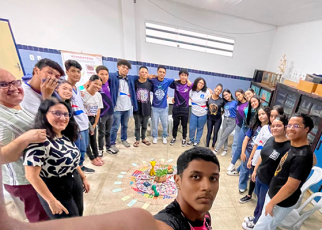 A imagem mostra um grupo de pessoas, entre jovens e adultos, formando um círculo em uma sala de aula. Todos estão sorrindo e abraçados, olhando para a câmera, que está sendo segurada por um dos jovens em primeiro plano. No centro do círculo, há um arranjo colorido no chão, com flores, objetos decorativos e cartões dispostos em círculos, formando uma mandala. O ambiente é claro, com paredes azuis e armários com livros ao fundo. A imagem transmite união e acolhimento.