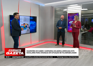rês apresentadores estão em um estúdio de televisão com iluminação clara e detalhes em vermelho. Eles sorriem e conversam enquanto uma tela exibe a imagem da campanha "Guardiões do Saber", que arrecada kits escolares para crianças em situação de vulnerabilidade. No rodapé da tela, há a tarja do programa Boletim Gazeta com a manchete da campanha e um pequeno noticiário rolando. Ao fundo, vê-se parte da redação do jornal.