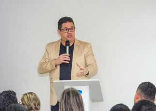Homem alto de cabelo preto em pé proferindo palestra com um microfone na mão direita e vestindo blusa preta e blazer creme