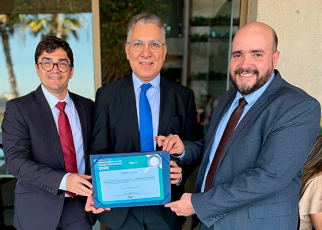 Há três homens trajados de terno e gravata, dois deles usam óculos. Eles estão sorridentes e seguram um certificado.