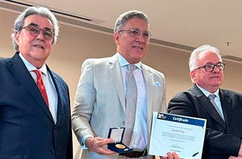 Três homens, sendo dois de terno escuro e um de terno claro segurando um certificado