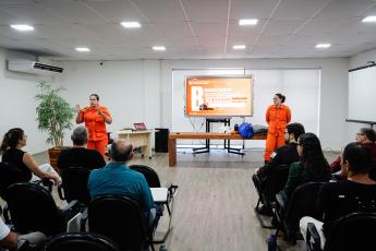Duas mulheres de macacão laranja ministrando aula para uma plateia formada por homens e mulheres, sendo uma do lado direito e a outra do lada esquerdo