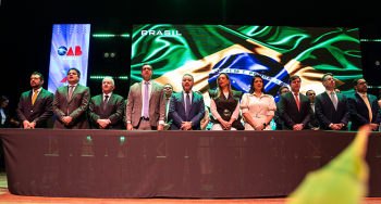 Homens e mulheres de pé em posse solene em frente a um telão com a imagem da bandeira do Brasil