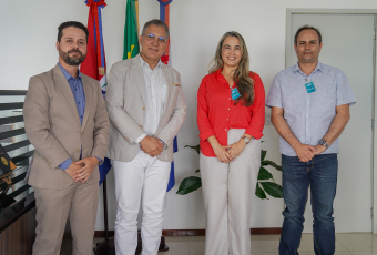 Na imagem, quatro pessoas estão em pé, lado a lado, posando para a foto em um ambiente interno. Atrás delas, há bandeiras e uma parede branca. Duas pessoas estão de terno, uma veste camisa social clara e calça jeans, e outra usa blusa vermelha e calça clara. Todos estão sorrindo.