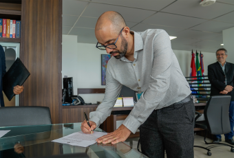 Na foto, um homem de óculos e barba assina um documento sobre uma mesa de vidro em um escritório moderno. Ele veste camisa social clara e calça escura, demonstrando formalidade. Ao fundo, é possível ver estantes com livros, mesas de trabalho e bandeiras posicionadas em um suporte, além de outras pessoas acompanhando o ato. A cena transmite solenidade e oficialidade, sugerindo a assinatura de um termo ou posse em cargo institucional.