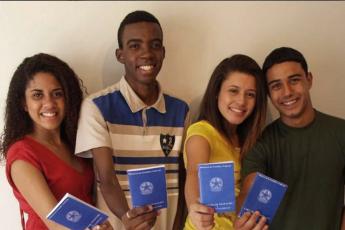 A imagem mostra quatro jovens sorridentes, lado a lado, segurando e exibindo suas carteiras de trabalho (CTPS – Carteira de Trabalho e Previdência Social), de cor azul. Eles parecem orgulhosos e felizes, possivelmente celebrando a conquista de uma oportunidade de emprego formal ou ingresso em um programa como o de Jovem Aprendiz. O fundo é neutro, o que mantém o foco nas pessoas e nos documentos que seguram.