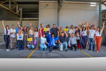 Várias pessoas, entre homens e mulheres, em pé de frente para a foto. Todos estão usando roupas leves e acenando com as mãos levantadas. Alguns estão à frente agachados