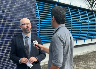 Um homem de terno e gravata está sendo entrevistado por um repórter que segura um microfone. Eles estão em área externa, em frente a uma parede de azulejos azuis e estruturas metálicas na mesma cor.
