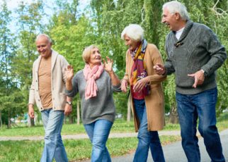 Há  quatro pessoas idosas caminhando juntas em um parque. Elas estão sorrindo, conversando e parecem se divertir. Estão vestidas com roupas de clima ameno.Ao fundo, há muitas árvores e vegetação, transmitindo um ambiente agradável e tranquilo.
