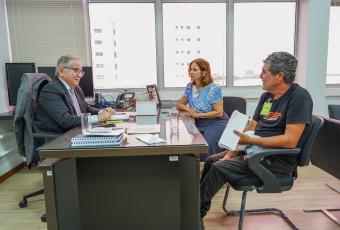 Três pessoa sentada ao redor de uma mesa, dois homens e uma mulher; há um homem de terno escuro que aparentemente está sentado do lado principal atendo o outro homem a a mulher, que vestem roupas informais