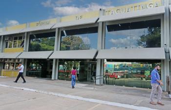 A foto mostra a fachada do Fórum de Taguatinga, no Distrito Federal. O prédio é de dois andares, com revestimento em pastilhas amarelas na parte superior e grandes janelas de vidro espelhado na parte central, que refletem árvores e o céu. Na parte térrea, há portas de vidro, uma delas sinalizada como “entrada” e outra como “saída”. Em frente ao fórum, algumas pessoas caminham: um homem de camisa branca à esquerda, uma mulher de blusa rosa e calça jeans entrando no prédio pelo centro, e outro homem de camisa