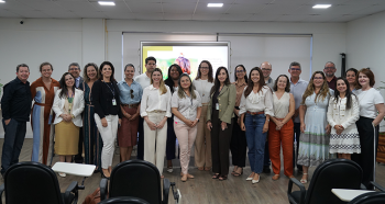 A imagem mostra um grupo de pessoas posando para uma foto em um ambiente interno, possivelmente uma sala de treinamento ou auditório. Elas estão organizadas em uma única fileira, sorrindo para a câmera. Ao fundo, há um telão ligado exibindo uma apresentação com texto e imagem, sugerindo que o grupo participou de uma atividade ou evento institucional. O ambiente é bem iluminado, com piso de madeira e cadeiras de cor escura dispostas à frente do grupo.