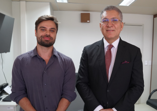 Há duas pessoas em pé, lado a lado.  À esquerda, há um homem com barba curta, cabelo castanho e camisa social roxa, com expressão serena. À direita, há um homem de terno preto, camisa branca e gravata bordô, usando óculos. Ele está com as mãos cruzadas à frente do corpo e sorri levemente. 