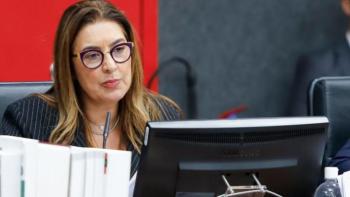 Mulher branca de cabelos loiros usando óculos e sentada; à sua frente, tem um computador preto