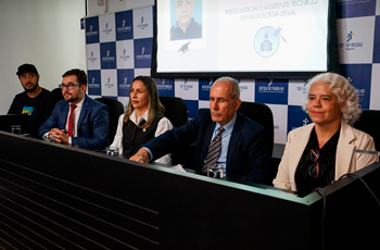 A imagem mostra uma mesa de autoridades durante uma solenidade. Cinco pessoas estão sentadas lado a lado, em um ambiente formal, com painel institucional ao fundo. Ao centro, um homem de terno e gravata fala ou ouve atentamente, ladeado por outros participantes — homens e mulheres — também trajando roupas formais. Um telão ao fundo exibe informações sobre o curso de “Perito Judicial e Assistente Técnico em Radiologia Legal”, indicando o contexto da formatura mencionada no texto.
