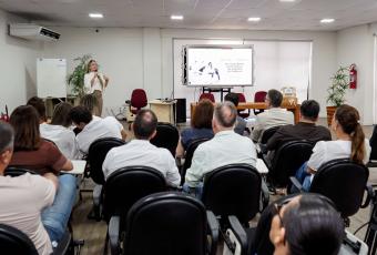 Imagem de uma palestra em uma sala de treinamento. À frente do ambiente, uma mulher em pé fala ao público com um microfone. Ao lado dela, uma tela exibe apresentação sobre a campanha Janeiro Branco, com foco em saúde mental no trabalho. À sua frente, há uma mesa com cadeiras vazias. O público, formado por homens e mulheres adultos, está sentado em fileiras de cadeiras, atento à palestra.