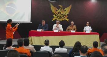 A imagem mostra uma mesa de autoridades em um auditório, com quatro representantes sentados à frente do público, durante um evento institucional. Ao fundo, há um grande brasão oficial na parede, e, à esquerda, uma pessoa em uniforme laranja registra o momento com o celular. A plateia acompanha a cerimônia sentada, em ambiente formal.