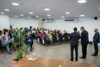 A imagem mostra uma sala de treinamento com plateia sentada em cadeiras organizadas em fileiras, assistindo a dois homens de terno que falam à frente do ambiente. O espaço é amplo, com iluminação em LED no teto, ar-condicionado nas paredes e uma câmera posicionada para registrar o evento. Em primeiro plano, há uma mesa de madeira com jarra e copos, além de uma planta decorativa.