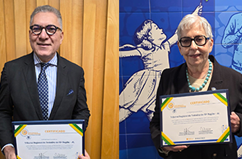 À esquerda, um homem de cabelos grisalhos usa óculos, terno escuro, camisa clara e gravata azul com pequenas estampas. Ele sorri levemente enquanto segura um certificado emoldurado com detalhes nas cores verde, amarelo e azul. Ao fundo, há uma cortina em tom amarelo-ouro.  À direita, uma mulher de cabelo curto e branco também usa óculos e veste um blazer escuro combinado com um colar de pedras em tom turquesa. Ela segura um certificado semelhante, igualmente emoldurado e com as mesmas cores na borda. 