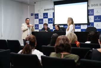 Homem fala ao microfone em auditório institucional, com equipe à mesa ao fundo e público sentado assistindo.