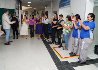 Há um grupo de muleres de pé aplaudindo.. À frente delas há um homem de terno claro. e calça zul. Ele fala para elas.