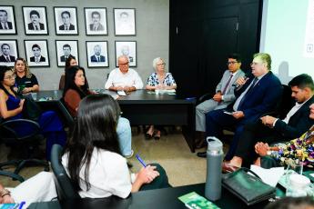 Reunião formal com várias pessoas sentadas; um homem de terno fala ao microfone enquanto os demais escutam.