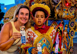 ulher adulta sorridente e criança lado a lado, ambos olhando para a câmera. A mulher segura um folheto informativo. A criança veste traje colorido e ornamentado típico de festa popular, com chapéu e fitas. Ao fundo, há decoração rica em detalhes e cores vibrantes, lembrando um cenário cultural ou festivo.