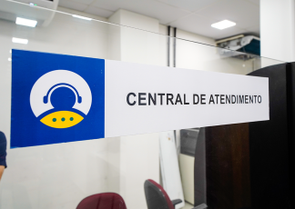 Placa de identificação com a inscrição “Central de Atendimento” afixada em uma divisória de vidro. À esquerda, há um ícone estilizado de uma pessoa com fone de ouvido, simbolizando suporte ao público. O ambiente ao fundo é interno, com iluminação artificial e aparência de escritório. É possível ver cadeiras e parte de uma mesa, indicando área de atendimento. A imagem transmite organização e foco em serviço ao usuário