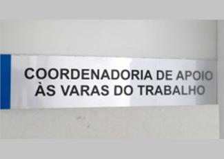 Foto de uma  placa afixada em uma porta cinza com o nome: Coordenadoria de Apoio às Varas do Trabalho escrito em letras pretas