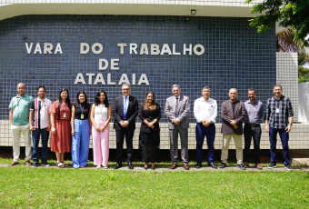 A imagem mostra um grupo de onze pessoas posando lado a lado em frente à fachada da Vara do Trabalho de Atalaia. Ao fundo, há uma parede revestida com pequenos azulejos azuis, onde se lê, em letras grandes e claras: “VARA DO TRABALHO DE ATALAIA”.  Os integrantes do grupo estão vestidos de forma formal e semiformal, com trajes que variam entre ternos, camisas sociais e roupas mais leves, sugerindo um ambiente institucional, mas também descontraído. Eles estão alinhados sobre uma área de grama.