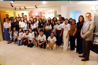 Um grupo numeroso de estudantes, acompanhado por alguns adultos, posa para uma foto em um espaço expositivo interno. Os jovens, em sua maioria usando camisetas padronizadas, estão organizados em fileiras, alguns agachados na frente e outros em pé atrás. Ao fundo, há painéis informativos sobre temas históricos, e o ambiente é bem iluminado, com aparência de museu ou memorial.