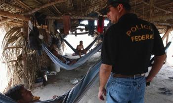 Um agente da Polícia Federal realiza fiscalização em um abrigo improvisado de trabalhadores, construído com madeira e palha. No interior do local, algumas pessoas descansam em redes penduradas sob a estrutura rústica. A imagem retrata condições precárias de alojamento em área de trabalho rural.