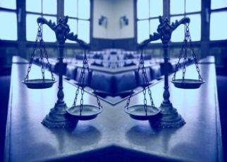 Foto de duas balanças da justiça em cima de uma mesa; ao fundo, janelas de vidros