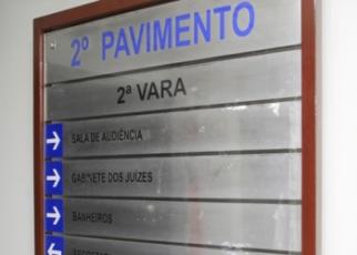 A imagem mostra uma placa de sinalização interna instalada em uma parede, indicando os setores localizados no 2º pavimento da 2ª Vara. No painel, aparecem direções para a Sala de Audiência, Gabinete dos Juízes e Banheiros, acompanhadas por setas indicativas. A placa possui fundo metálico, moldura em tom amadeirado e inscrições em azul e preto, características comuns de ambientes institucionais e administrativos.